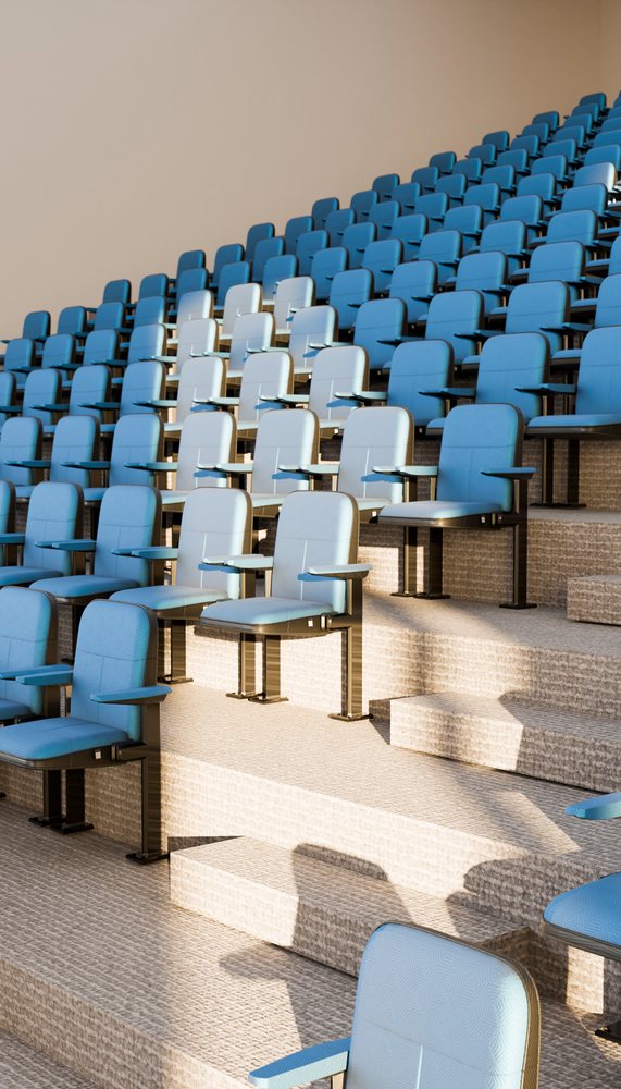 Lecture hall with tiered blue cushioned chairs and built-in desks in a modern educational interior with bright natural lighting.