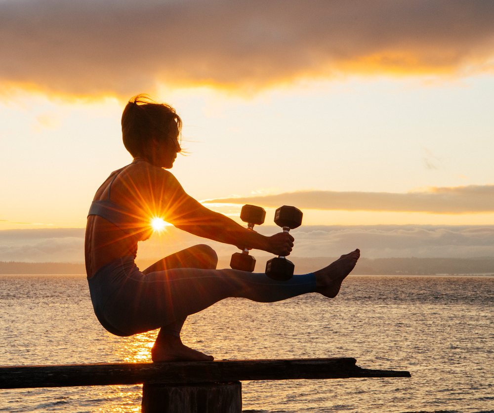 Woman balancing barefoot on a wooden beam over calm water at sunset while holding dumbbells, WA, USA.