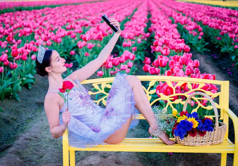 Ballet dancer in tutu and tiara takes a selfie sitting barefoot on a yellow bench amid pink tulip fields, USA.