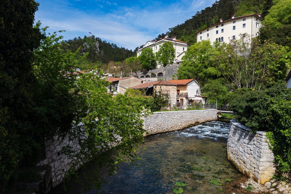 The Vipava River flows through a scenic Slovenian town with houses, stone walls, dense greenery, and a flag-topped hill under a clear blue sky, landscape, cityscape, Slovenia.