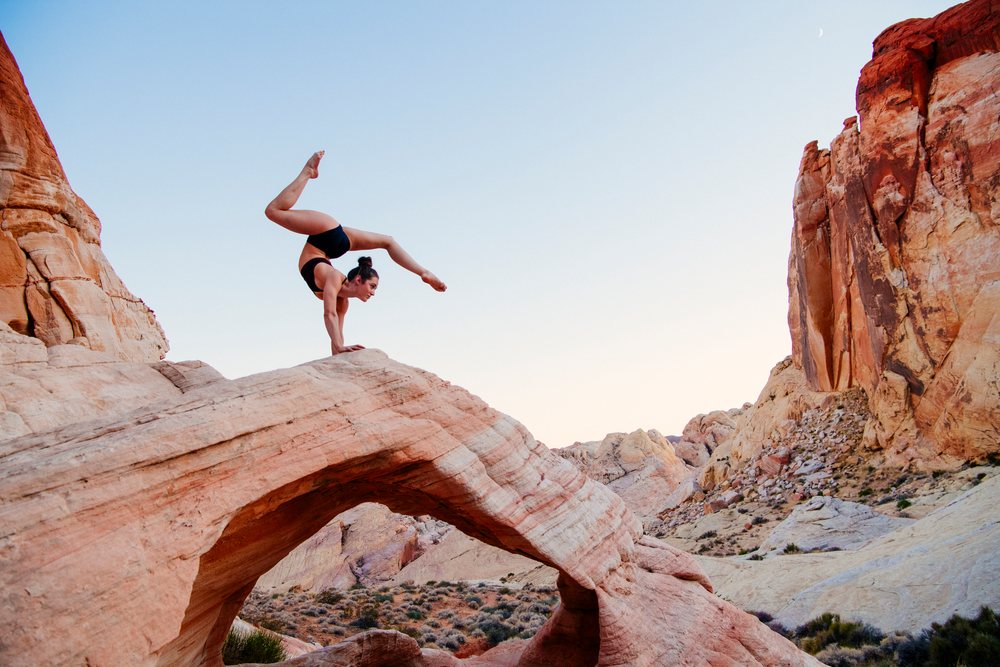 Woman in athletic wear performs a yoga handstand on a natural arch rock formation in the desert at sunset, USA.
