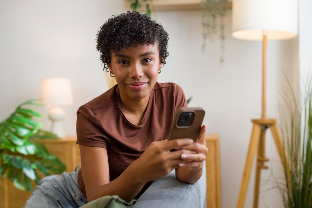 Young woman with short curly hair using a smartphone while relaxing in a casual home interior with modern decor and greenery.