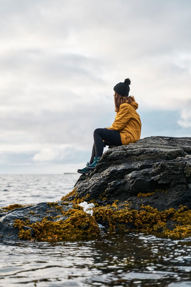 A white woman sits on a rocky coast by the ocean