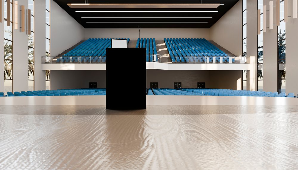 Podium with microphone on stage facing blue auditorium seats in a modern lecture hall with large windows and wood floor.