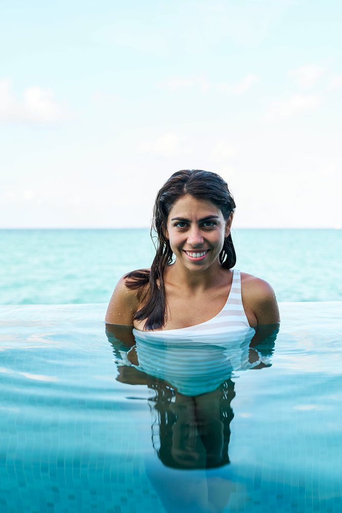 A woman wearing a white swimsuit is standing and smiling in an infinity swimming pool with the clear blue sea in the background in the, Maldives.