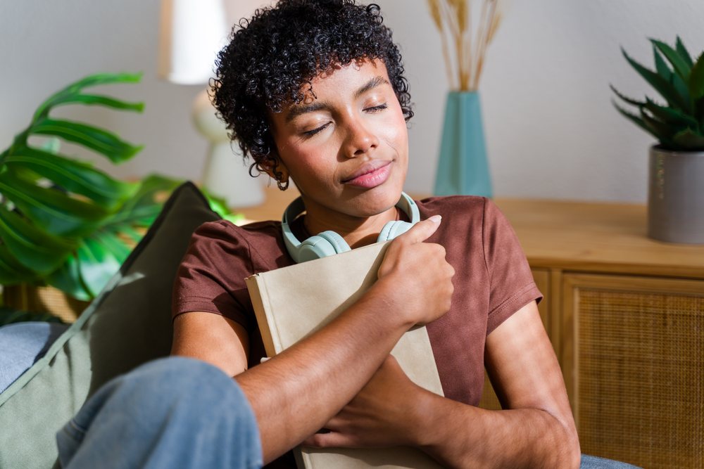 Woman sits indoors on a couch hugging a book with closed eyes and headphones around her neck, Spain.