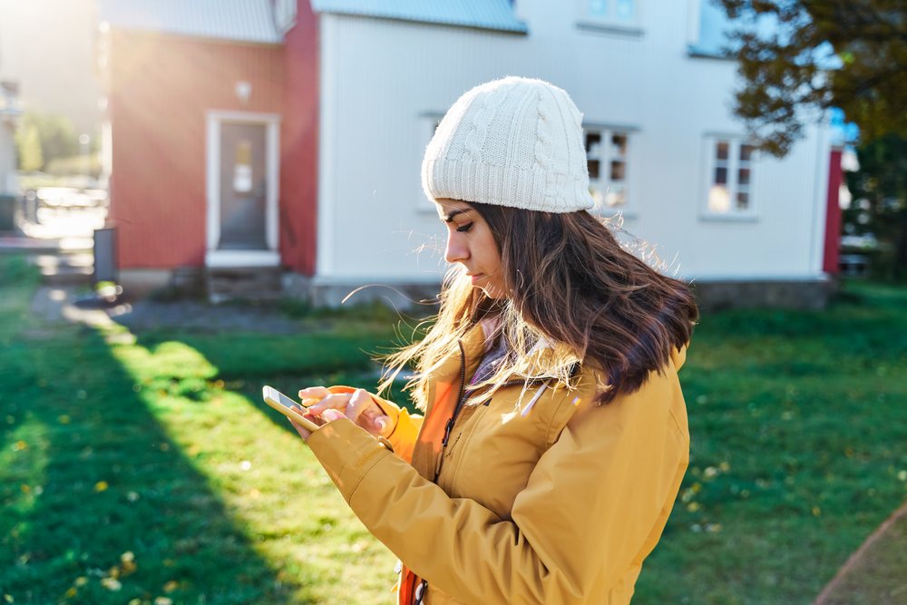 A woman wearing a yellow jacket and knit hat uses a smartphone outdoors on a sunny day with green grass, photographed, Iceland.