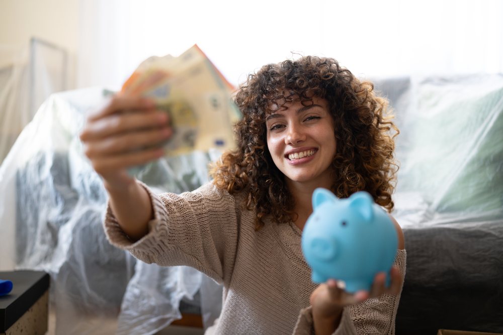 Smiling woman with curly hair holding Euro banknotes and a blue piggy bank in a living room with plastic covered furniture, moving, Spain.