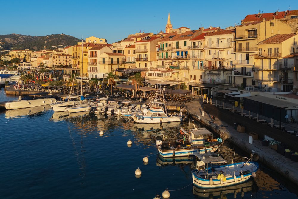 Boats moored at a harbor dock with Mediterranean-style buildings reflecting on the water during sunset, Travel Photography, Calvi, Corsica, France.