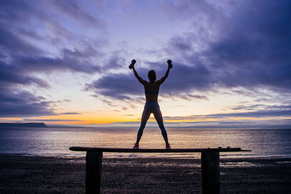 Woman exercising with dumbbells silhouetted on a beach at sunset, with ocean and sky in the background, WA, USA.