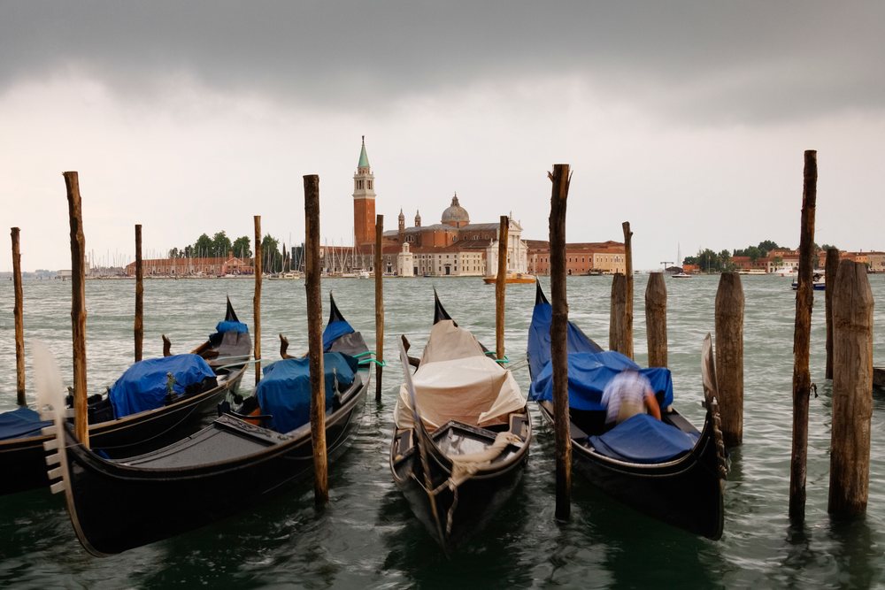 Gondolas moored to wooden poles on water with San Giorgio Maggiore church and surrounding buildings, Venice, Italy.