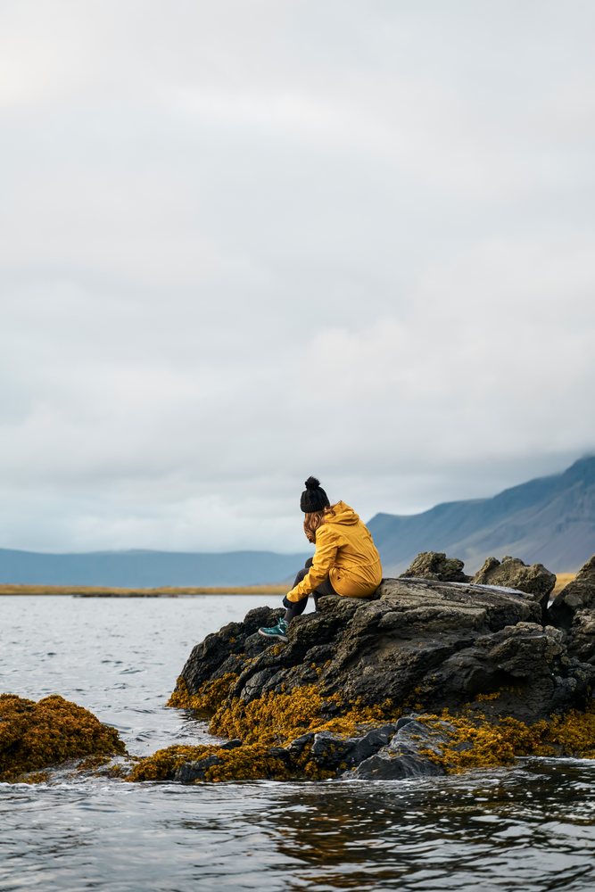 A white person sitting on seaweed-covered rocks by the sea