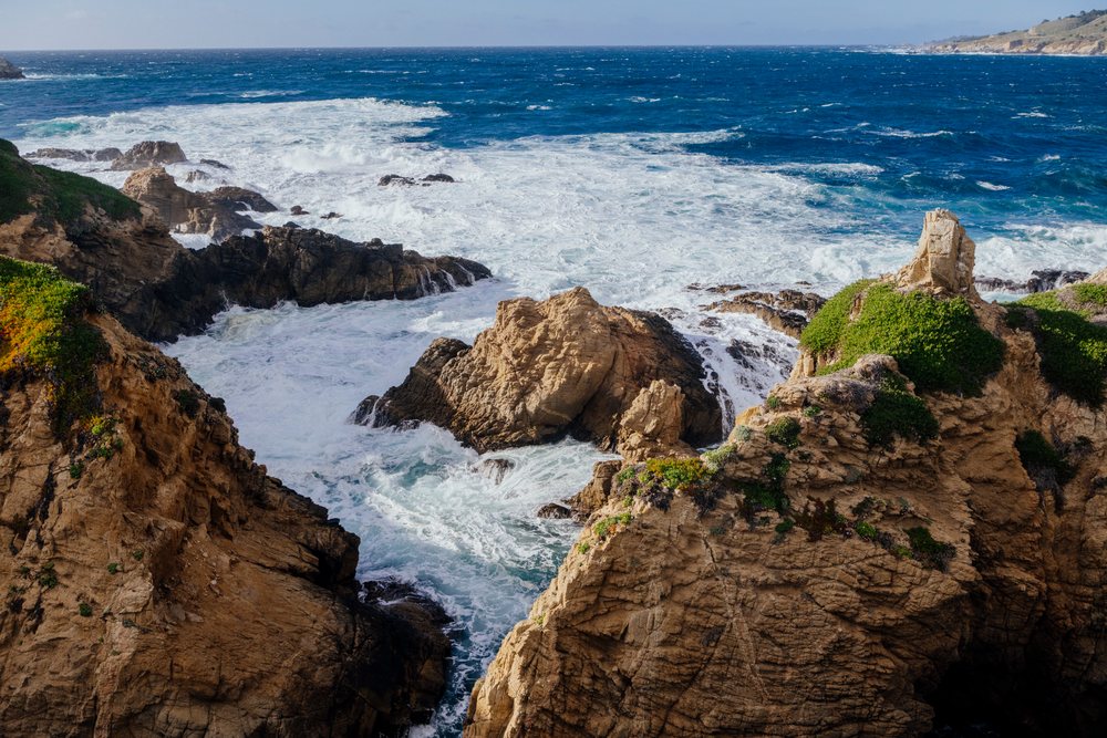 Waves crash against sea cliffs on a rocky coastline with green vegetation and blue Pacific Ocean water, California, USA.