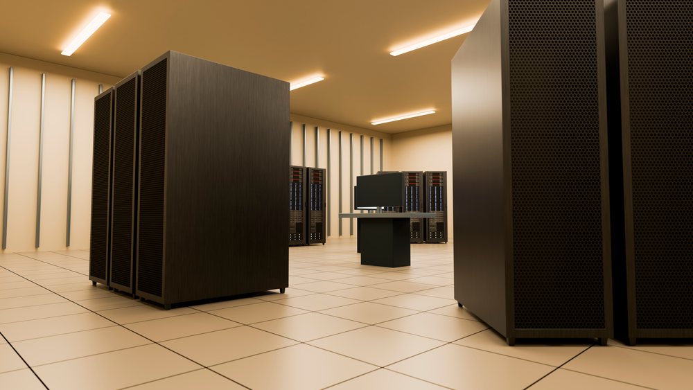 Flat screen monitor on table surrounded by server racks with ventilation panels inside a modern data center with tiled floor and ceiling lights.