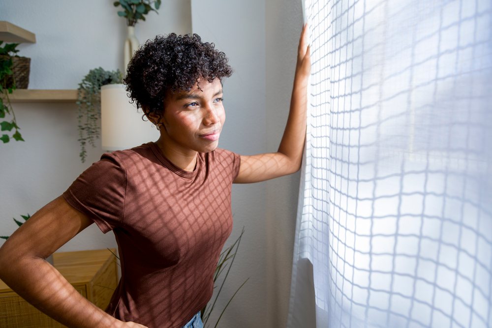 Young woman stands by a window curtain inside a home.