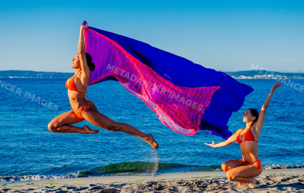 Two women perform partner yoga with flowing fabric on a beach