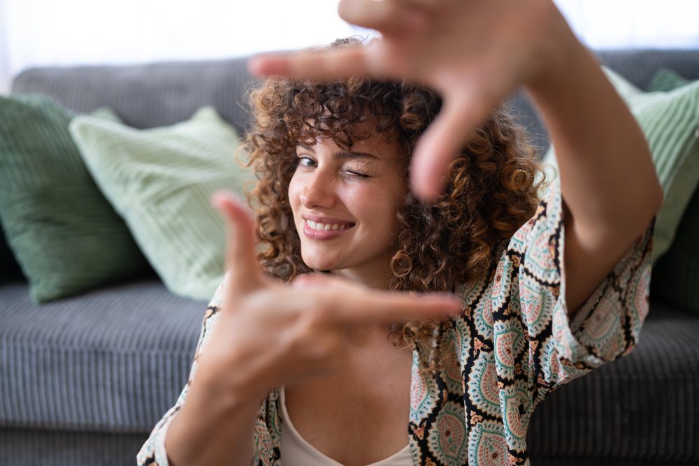 Woman with curly hair smiles and winks while framing her face with her hands indoors on a sofa with green cushions, home, girl, Spain.