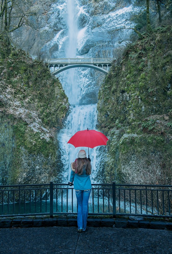 Woman holding a red umbrella stands behind a metal railing facing two-tier Multnomah Falls and a pedestrian bridge in rainy, cold weather, Bridal Veil, Oregon.