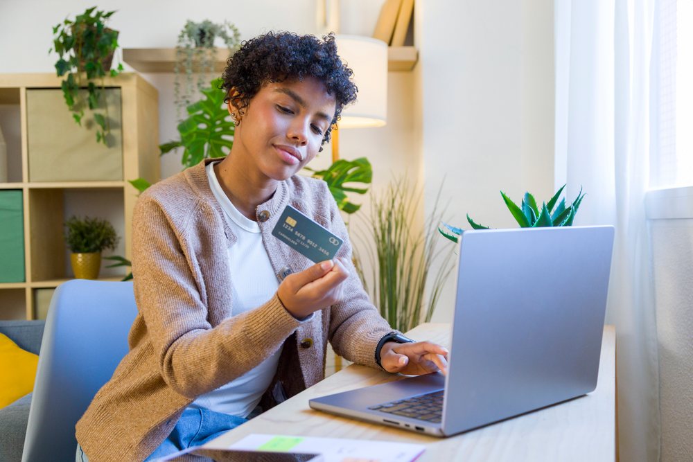 Woman seated at desk using credit card and typing on a laptop in a home office with indoor plants, remote work.