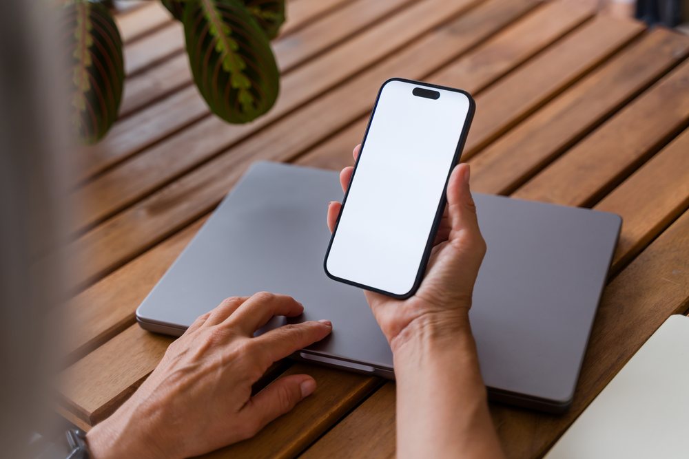 Hand holding a smartphone with a blank screen over a closed laptop on a wooden table near plant leaves, Spain.