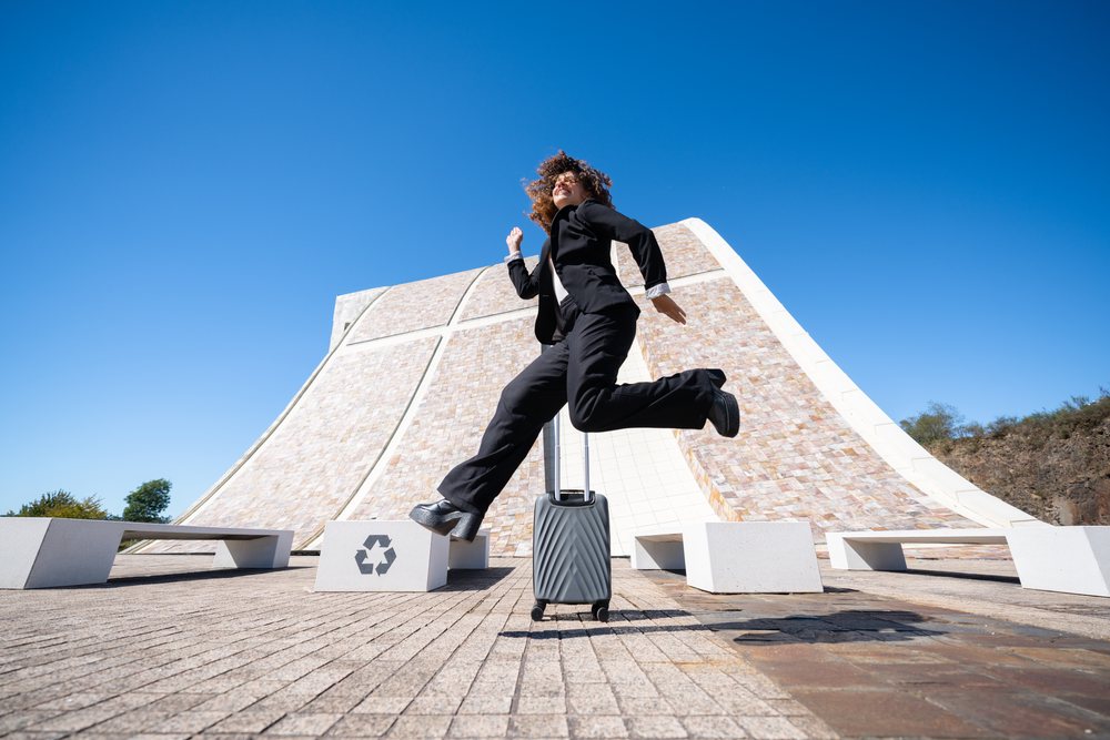 Female professional jumping mid-air with luggage near a modern curved building featuring recycling symbols on benches in Santiago de Compostela, businesswoman, traveling executive, Spain, Antiago De Compostela.