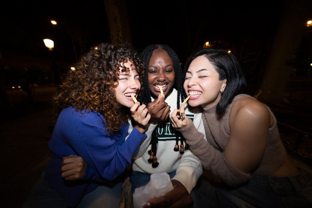 Three women laughing and eating french fries outdoors at night under street lamps, Madrid, Spain.