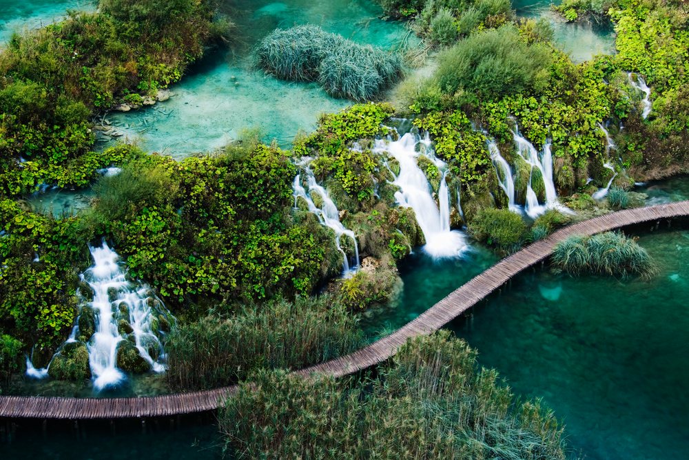 Multiple small waterfalls flow beside a wooden footbridge through dense green foliage, Plitvice Lakes National Park, Croatia.