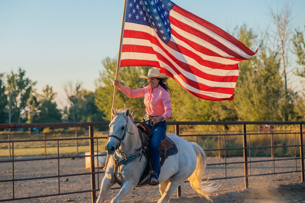 Woman riding a white horse holding an American flag at a ranch in the Southwest USA at sunset.