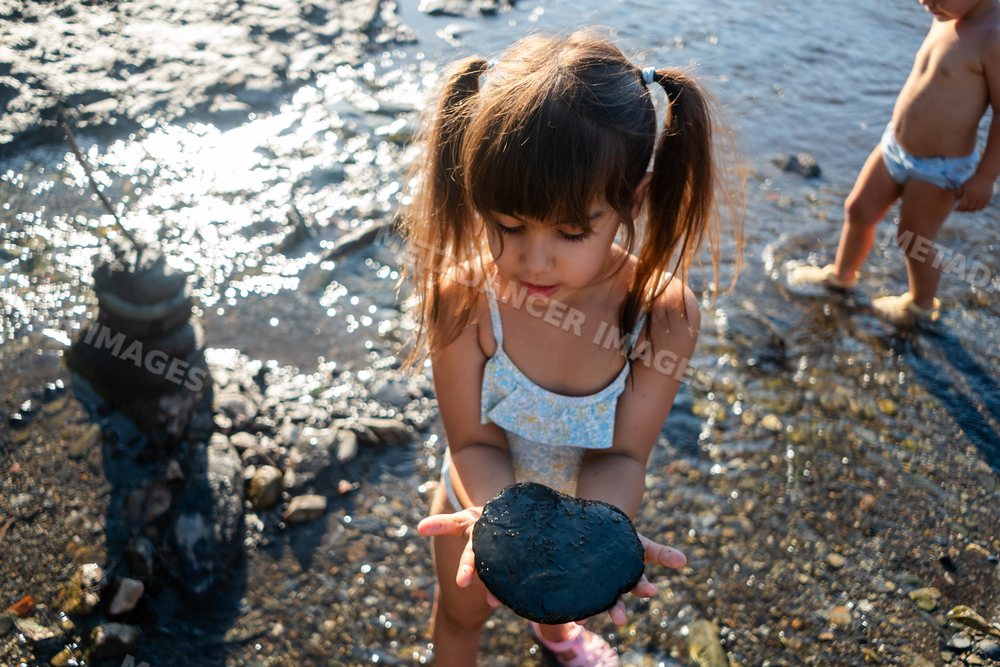 A young white girl holds a black rock by the water's edge