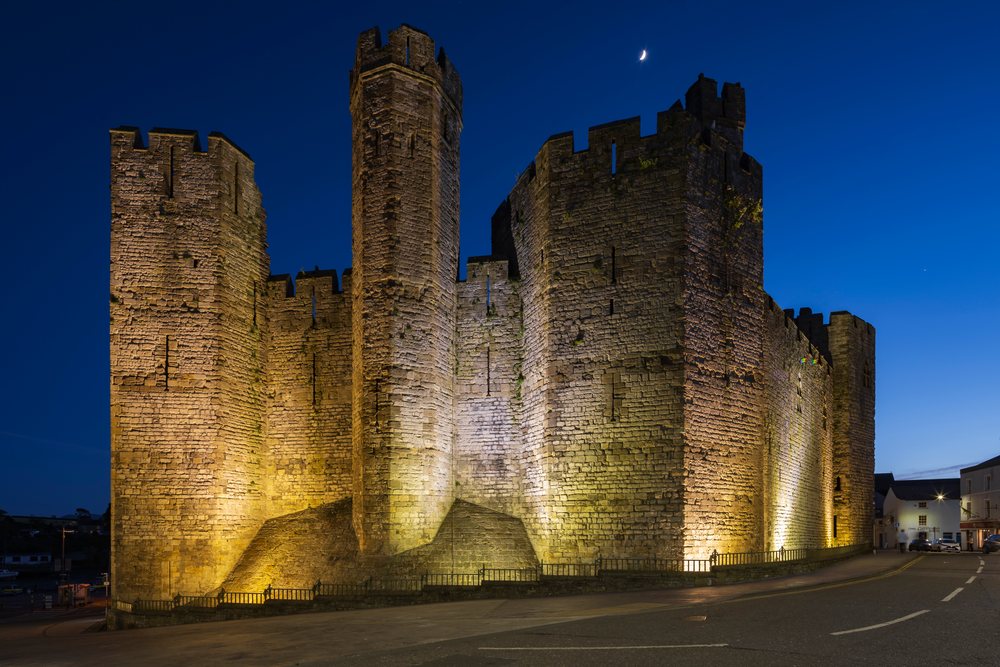 Stone castle walls and battlements of Caernarfon Castle are illuminated at dusk beneath a crescent moon, Landscape, Gwynedd, Wales.