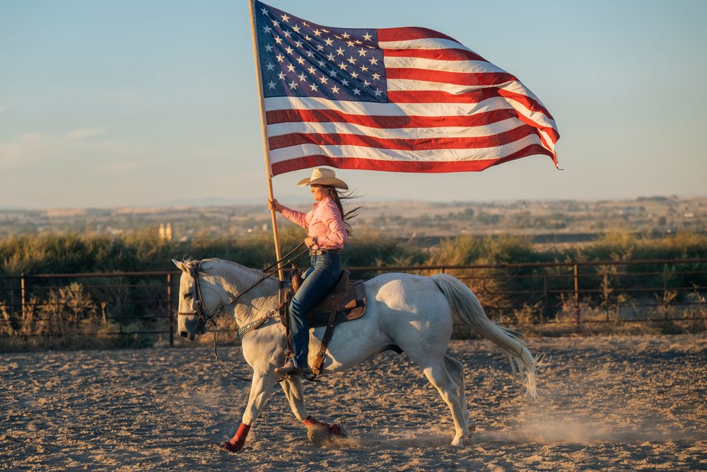 Woman riding a white horse holding a large American flag in a dusty outdoor arena at sunset, Southwest USA.