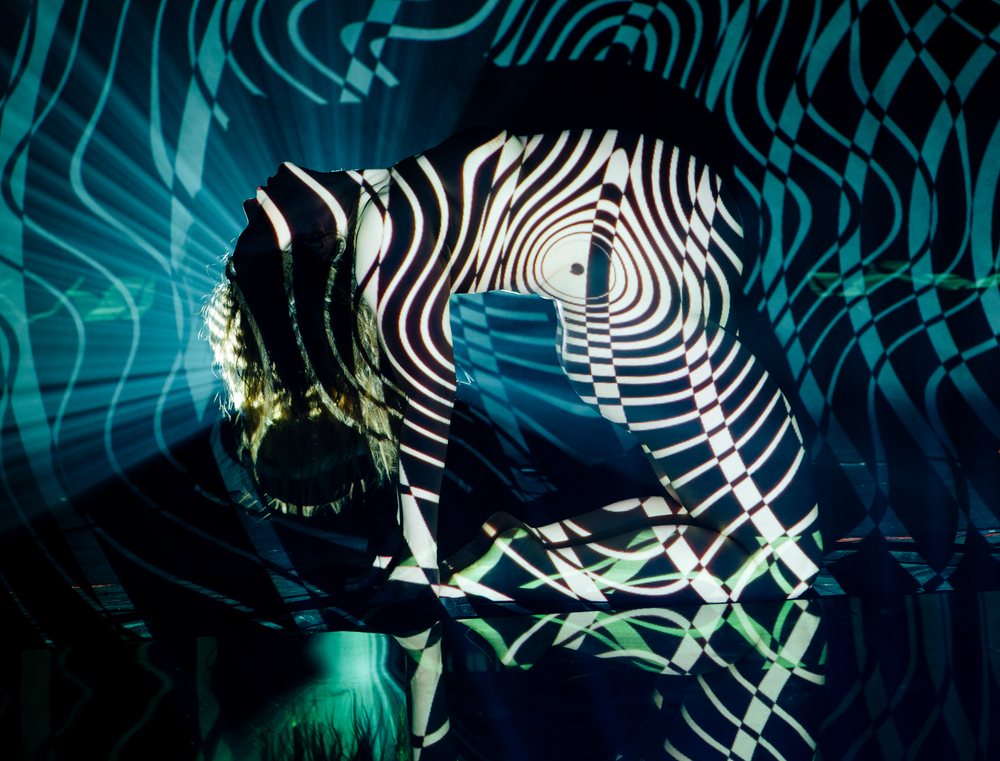 Woman kneeling with head tilted backwards under abstract striped light projection, with shadow and reflection in a dark studio, pregnancy.