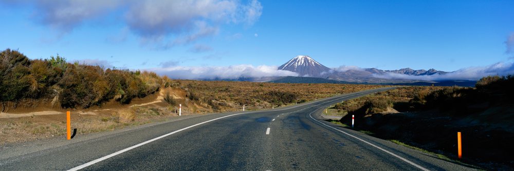Asphalt road leads through shrubland toward Mount Ngauruhoe with snow-capped peak under blue sky and clouds, New Zealand.