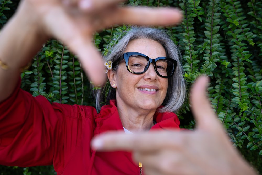 Woman with gray hair framing view with hands outdoors before green leaves, Spain.