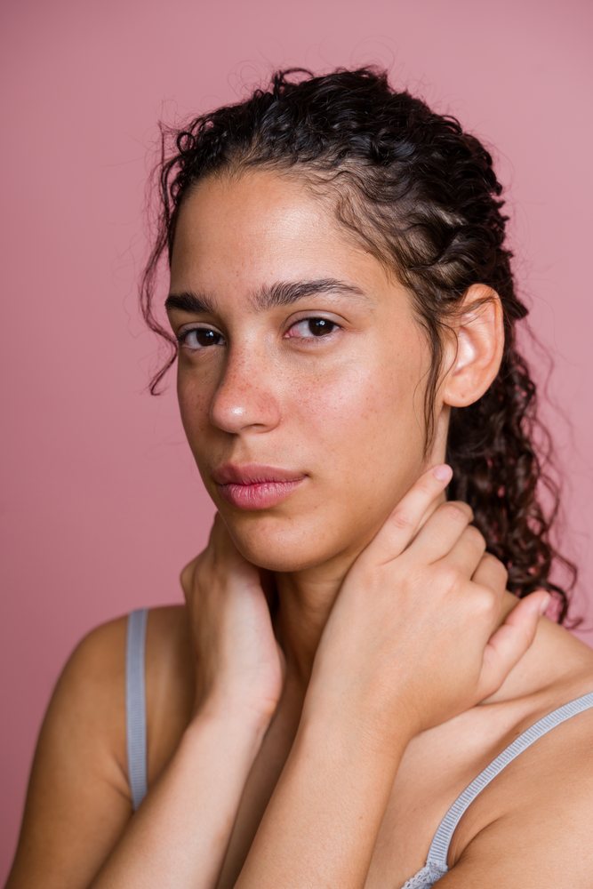 Woman with curly hair and natural skin poses with hands near face against a pink background in a studio setting, beauty shoot, Spain.