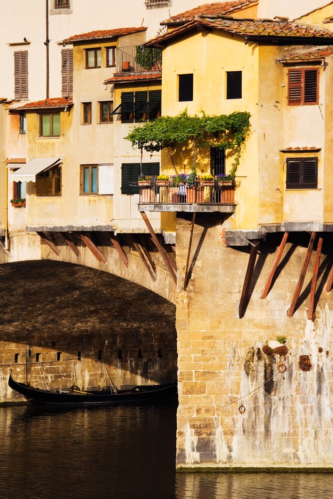 Ponte Vecchio historic stone arch bridge with yellow buildings and a black gondola reflecting on the river, Florence, Italy.