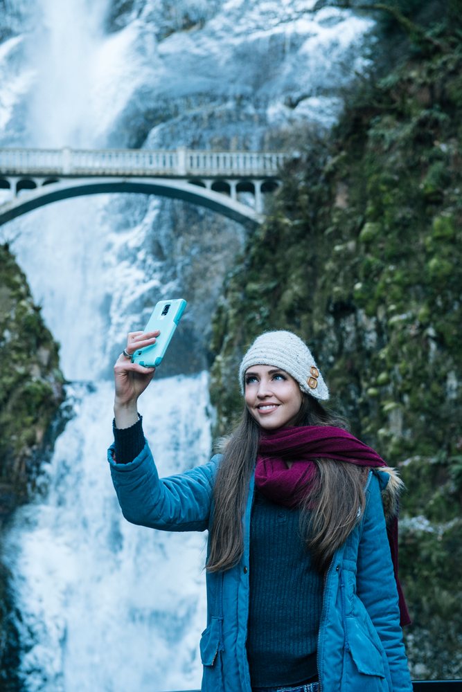 Smiling woman wearing winter clothing takes a selfie with a bridge and scenic natural landscape in the background, Multnomah Falls, Bridal Veil, Oregon.