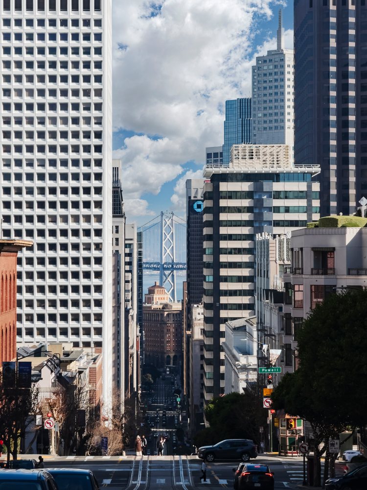 View down a San Francisco street with buildings, cars, pedestrians, and Bay Bridge in the background under a cloudy sky, landscape, cityscape, city life, California, USA.