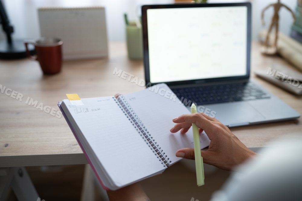 A white person writing in a notebook while using a laptop calendar