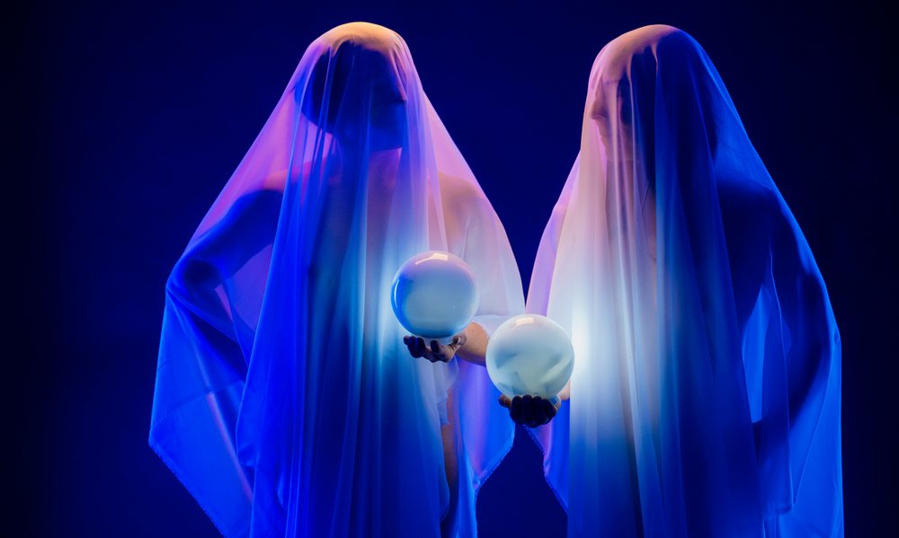 Two ghost figures covered in white sheets hold glowing crystal balls against a dark blue studio background with blue lighting, ghosts, USA.