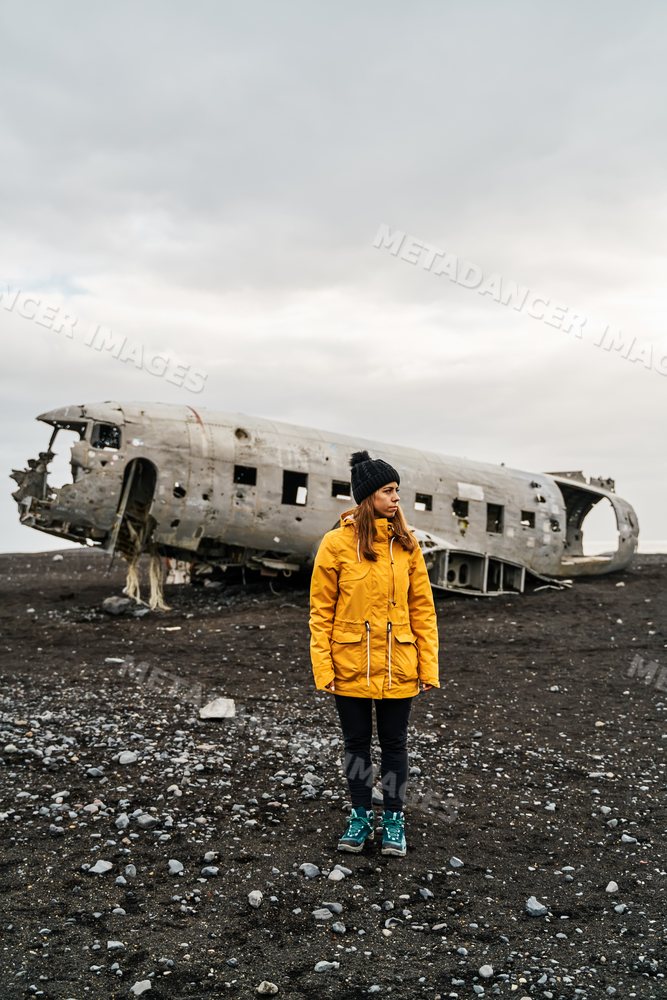 A white woman stands by a crashed airplane wreck on volcanic terrain