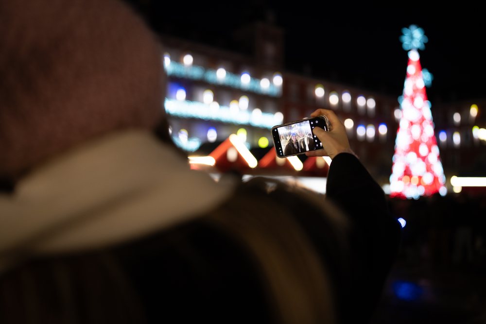 Two people take a selfie at a crowded outdoor Christmas market with a decorated tree and festive lights in Madrid at night, Spain.