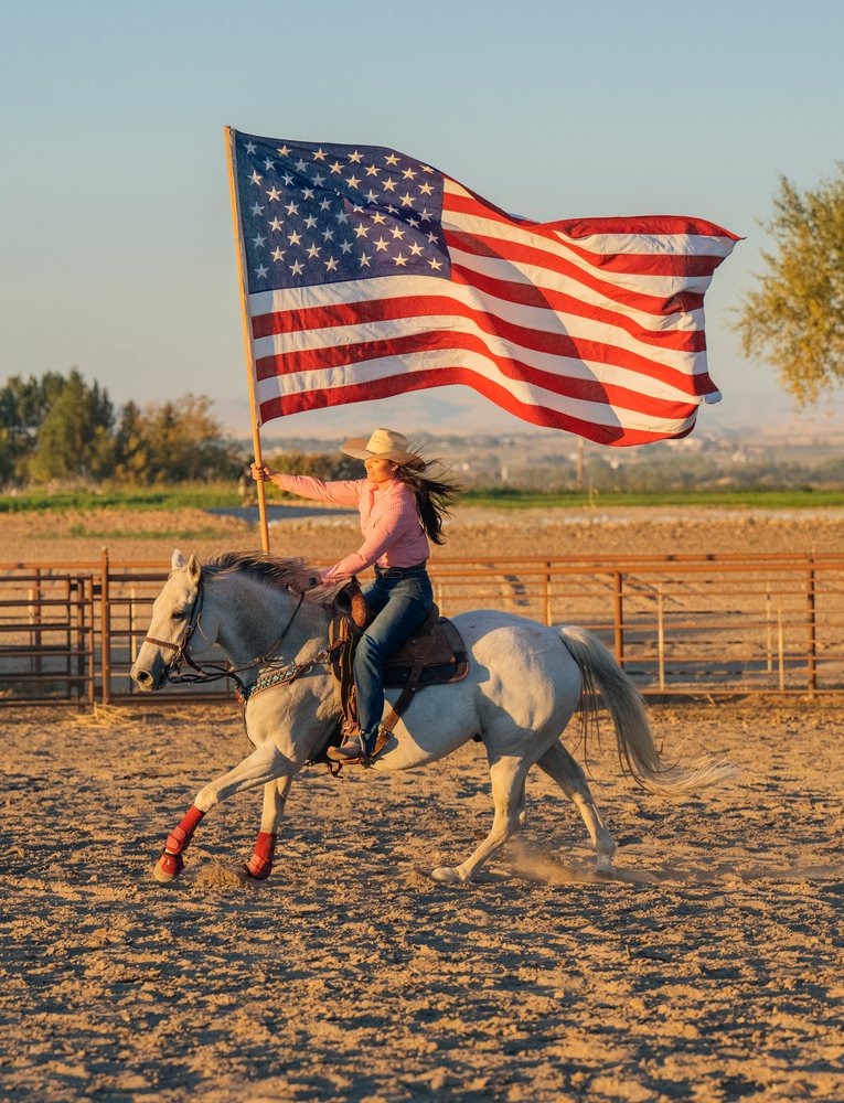 Woman wearing a cowboy hat rides a white horse with red leg wraps while holding a large American flag in a fenced outdoor arena, Southwest USA.
