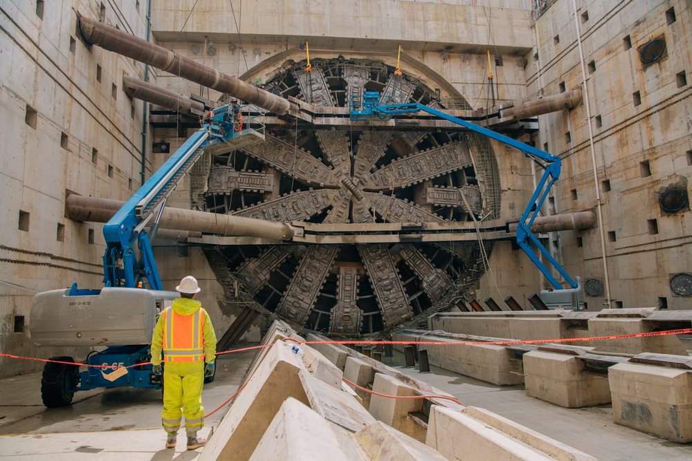 A construction worker in safety gear stands facing a tunnel boring machine inside an underground construction site, Bertha, drilling, USA.