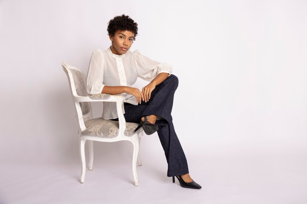 Woman seated on a white painted wooden chair wearing black high heels, black pants, and a white blouse in a studio with a white seamless background, fashion, young culture.