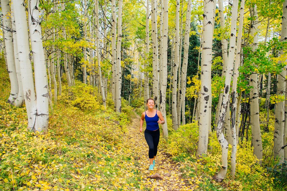 Woman running along a dirt trail through an aspen tree forest with yellow and green fall leaves, during daytime, Durango, Colorado, USA.