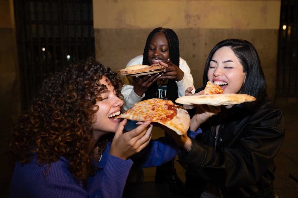 Three women eat cheese pizza slices together outdoors at night in a casual social setting, Madrid, Spain.