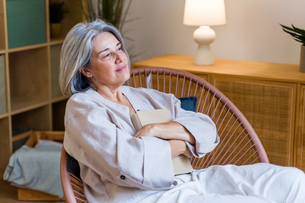 Mature woman with gray hair relaxes reading a book seated in a circular string chair inside a Spanish home, Spain.