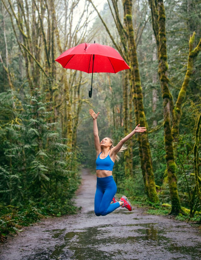 Woman in blue workout clothes jumping with a red umbrella on a wet forest path, USA.