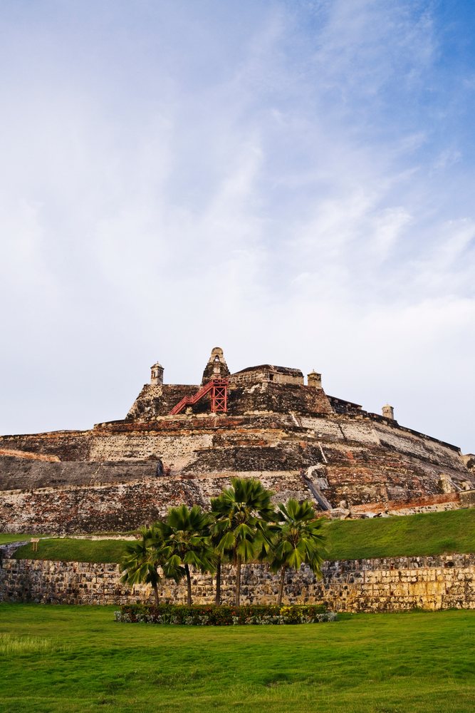 Castillo de San Felipe de Barajas fortress with stone and brick walls, palm trees, and green grass under a blue sky with clouds, Colombia.